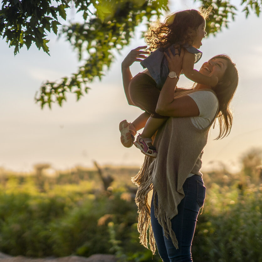 A woman holds her daughter up in the air and smiles. They are standing outside in front of a beautiful rural landscape at sunset.